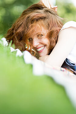 A smiling red-haired woman with a bright smile, lying on the grass with a white fence in the background.