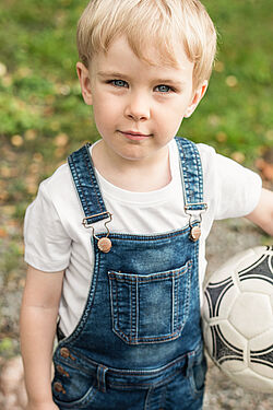 A young boy wearing denim overalls and a white shirt, holding a soccer ball, standing outdoors 