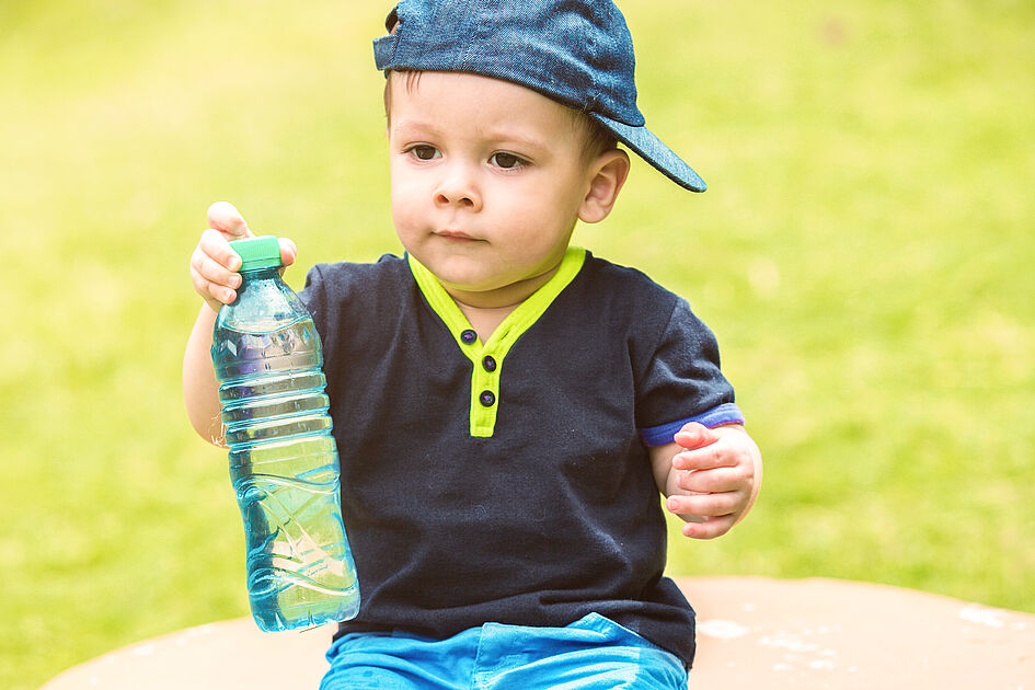 Young toddler wearing a cap and a navy shirt with neon green accents, holding a bottle of water while sitting outdoors on a sunny day.