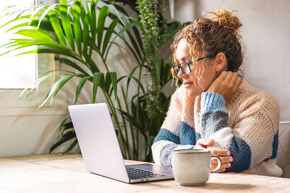 A woman with curly hair and glasses sitting at a table, looking at a laptop with a cup of coffee beside her.