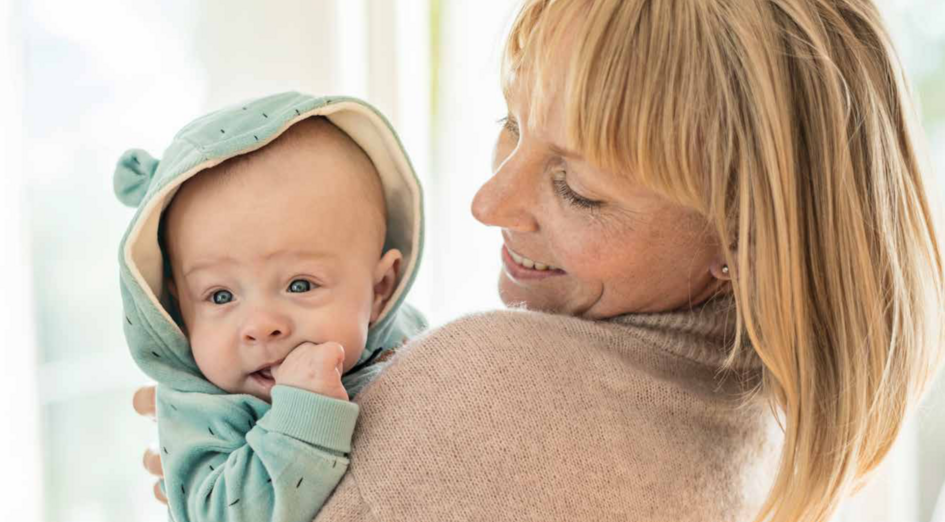 A woman holding a baby in a light blue hoodie, smiling at the baby while the baby looks into the camera, biting their hand