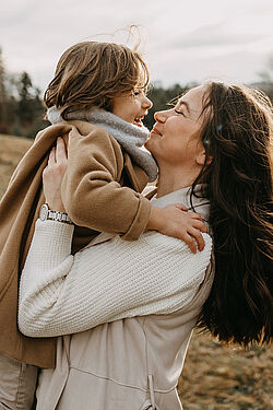 Mother holding and smiling at her child in an outdoor setting, both dressed warmly in coats and scarves on a cloudy day.