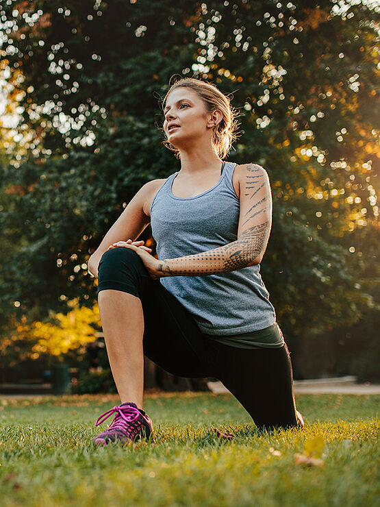 A woman in athletic wear stretches in a park with the sun shining through the trees.